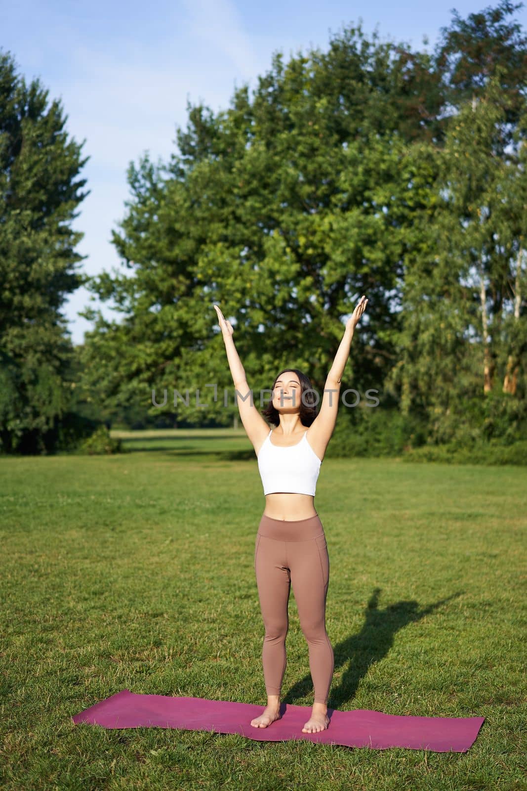 Vertical shot of asian woman raising hands up to sky, meditating, practice yoga, doing wellbeing training in park on green lawn.