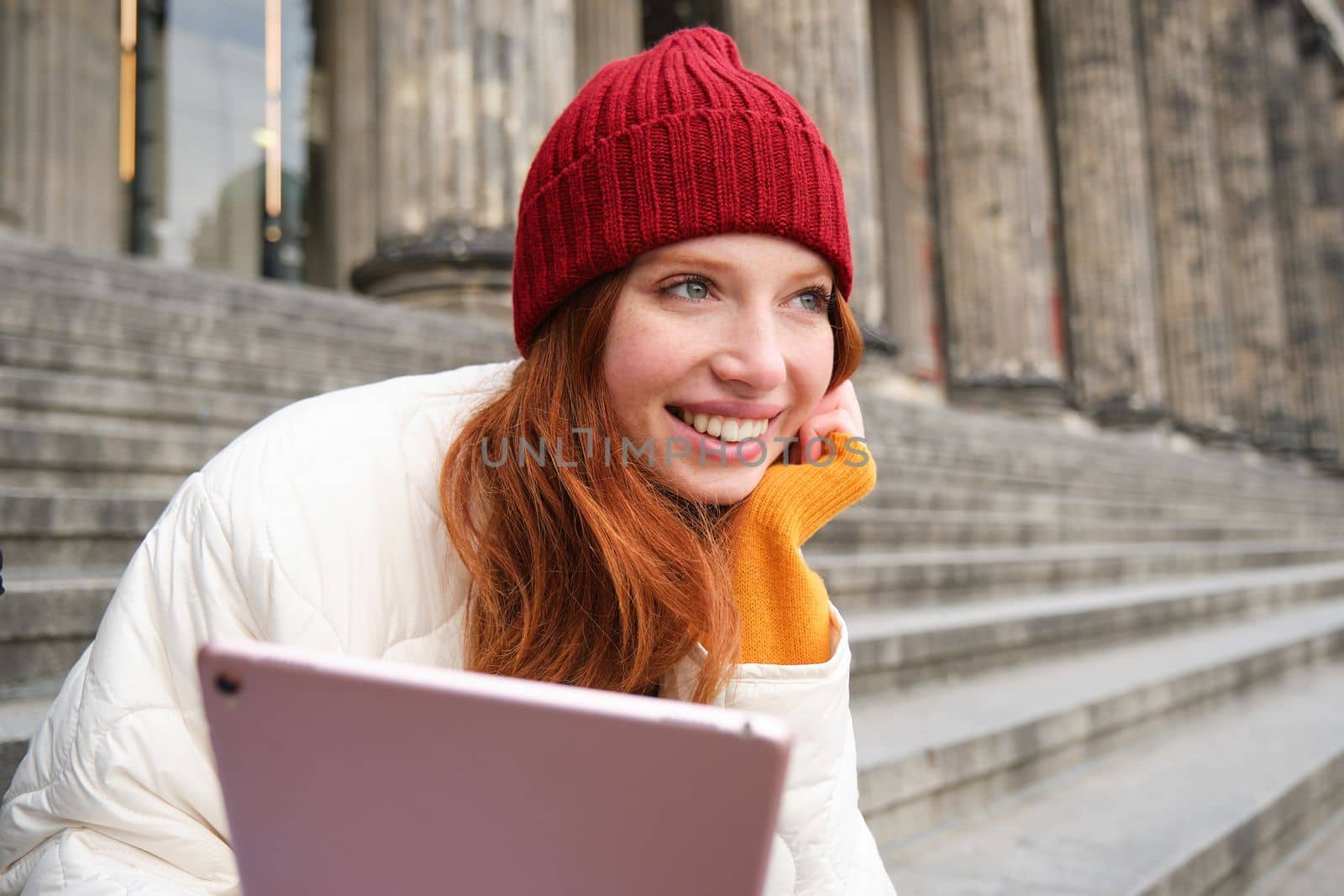 Outdoor shot of young stylish redhead girl sits on staircase and connects to public wifi, uses digital tablet, reads news on gadget by Benzoix
