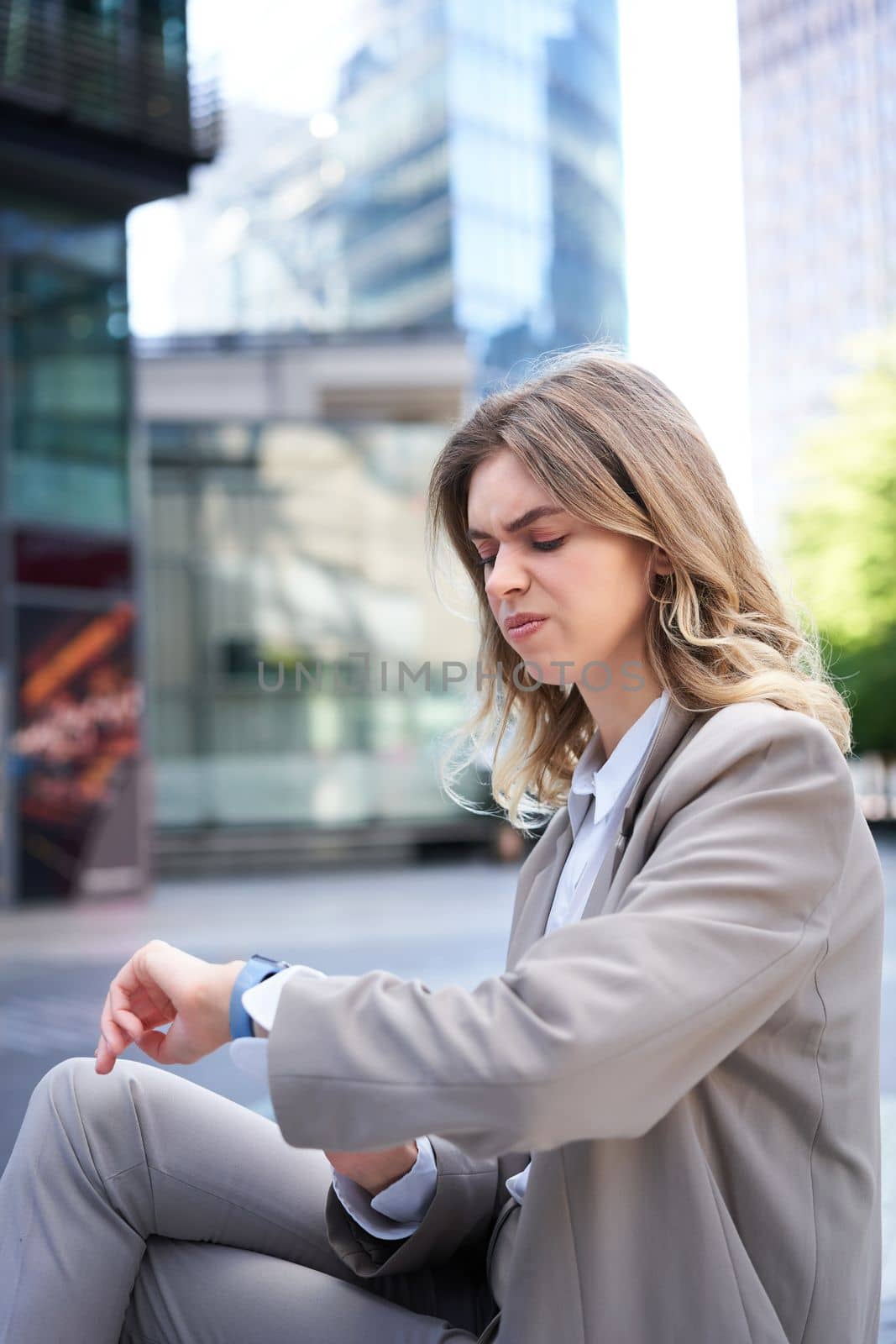 Portrait of office worker in suit, woman looking at watch with disappointed face, reading upsetting message, sitting outdoors by Benzoix