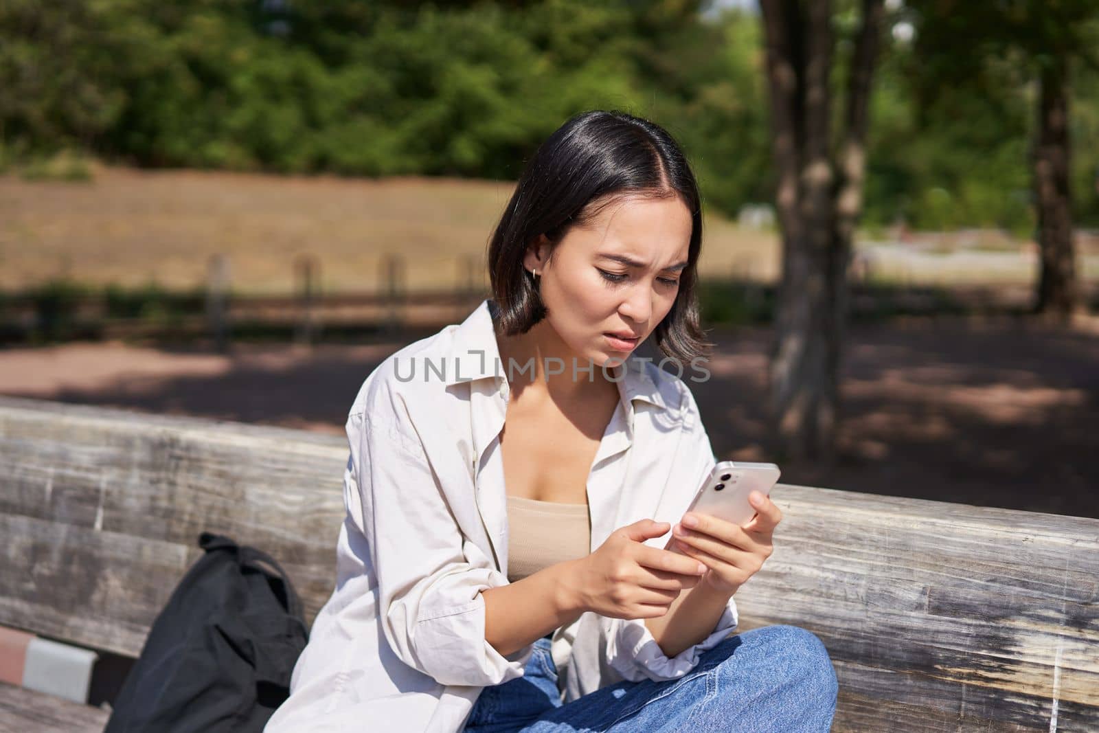 Asian girl frowning, looking concerned at her mobile phone, reading sad message, bad news on smartphone app, sitting on bench in park worrying.