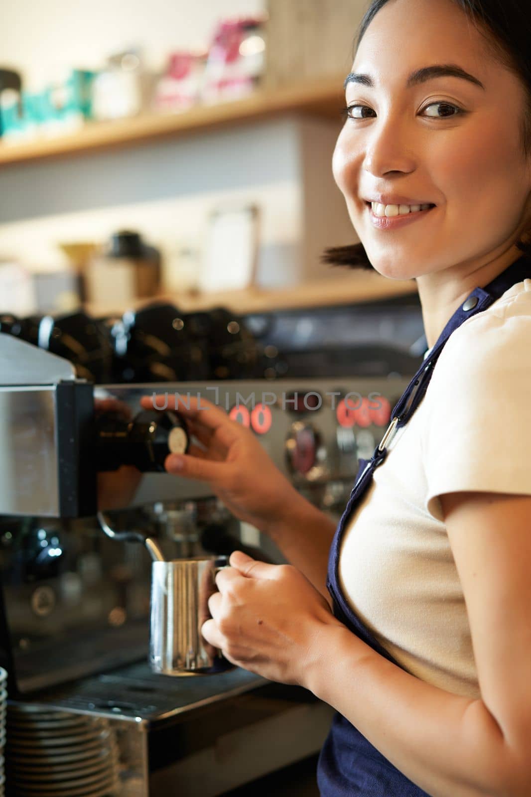 Vertical shot of barista, asian girl steaming milk for cappuccino, prepare latte for client, wearing blue apron, smiling happily, working in cafe.