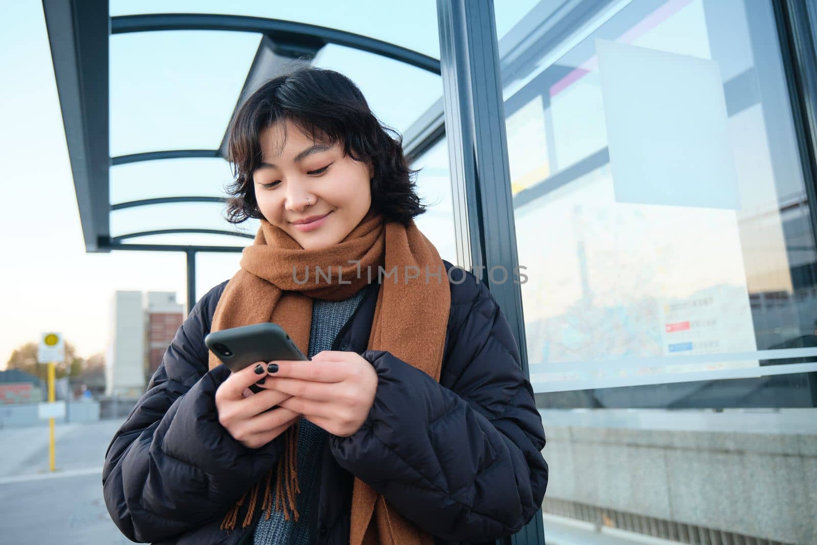 Beautiful Japanese girl, student standing on bus stop in winter jacket, holding smartphone, using application to track her public transport, ordering a ride by Benzoix