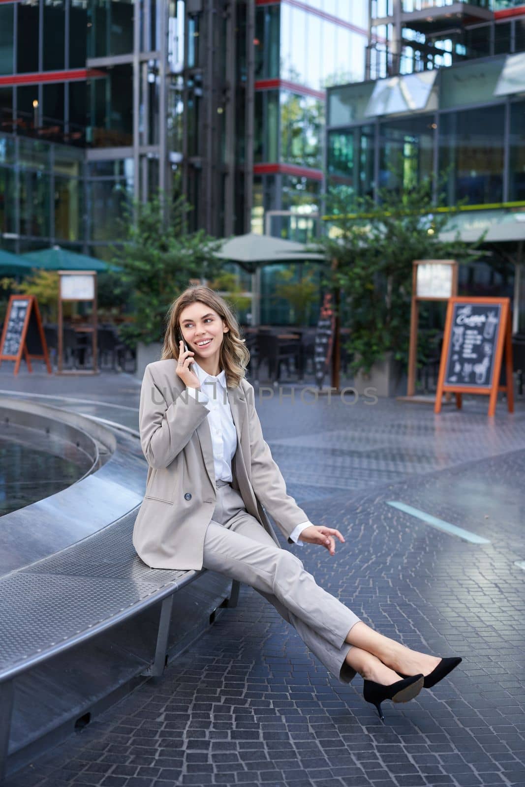 Vertical shot of young businesswoman in suit, sitting casually outside office buldings, talking on smartphone, making a phone call and smiling.