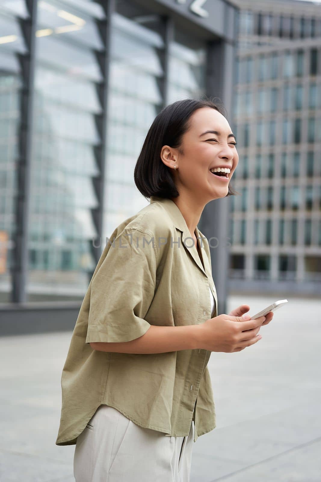 Vertical shot of beautiful asian woman walking on street, laughing and looking happy, enjoying the day.