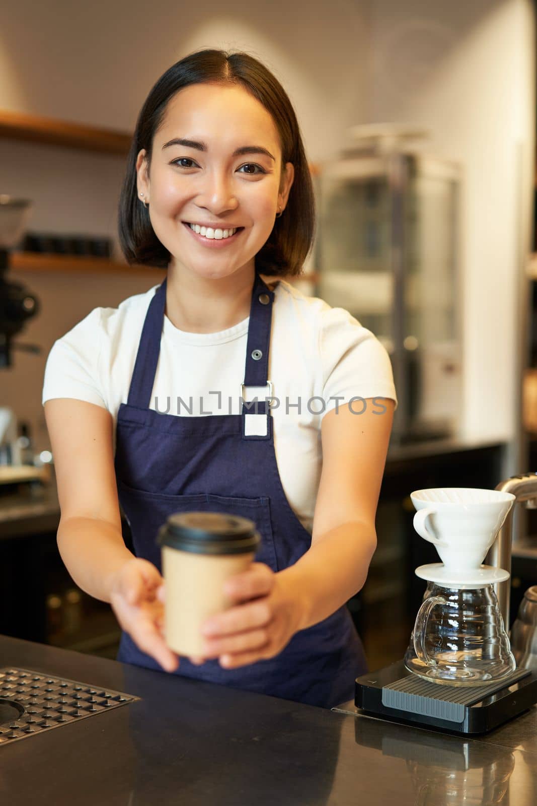 Smiling asian girl barista giving order to client, holding takeaway coffee cup, wearing apron, working behind counter in cafe.