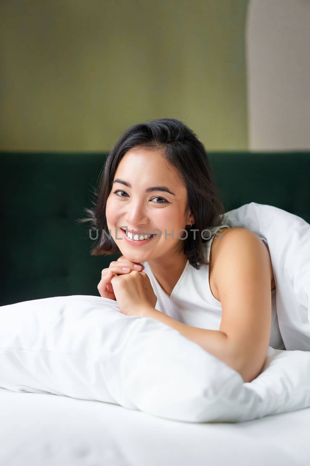 Vertical shot of smiling korean woman lying on pillow in her bed, spending morning in bedroom, waking up from sleep, looking happy.
