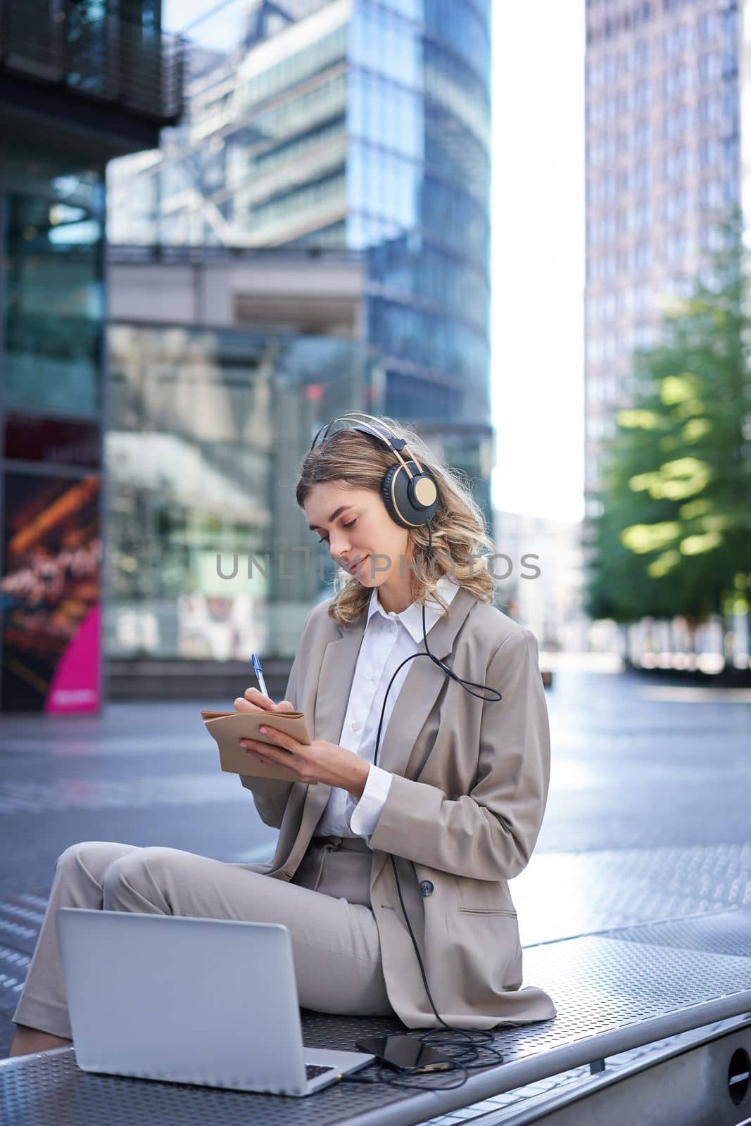 Vertical shot of smiling adult woman studying outdoors, listening to online course, taking notes, attend team meeting via app in laptop, using headphones.