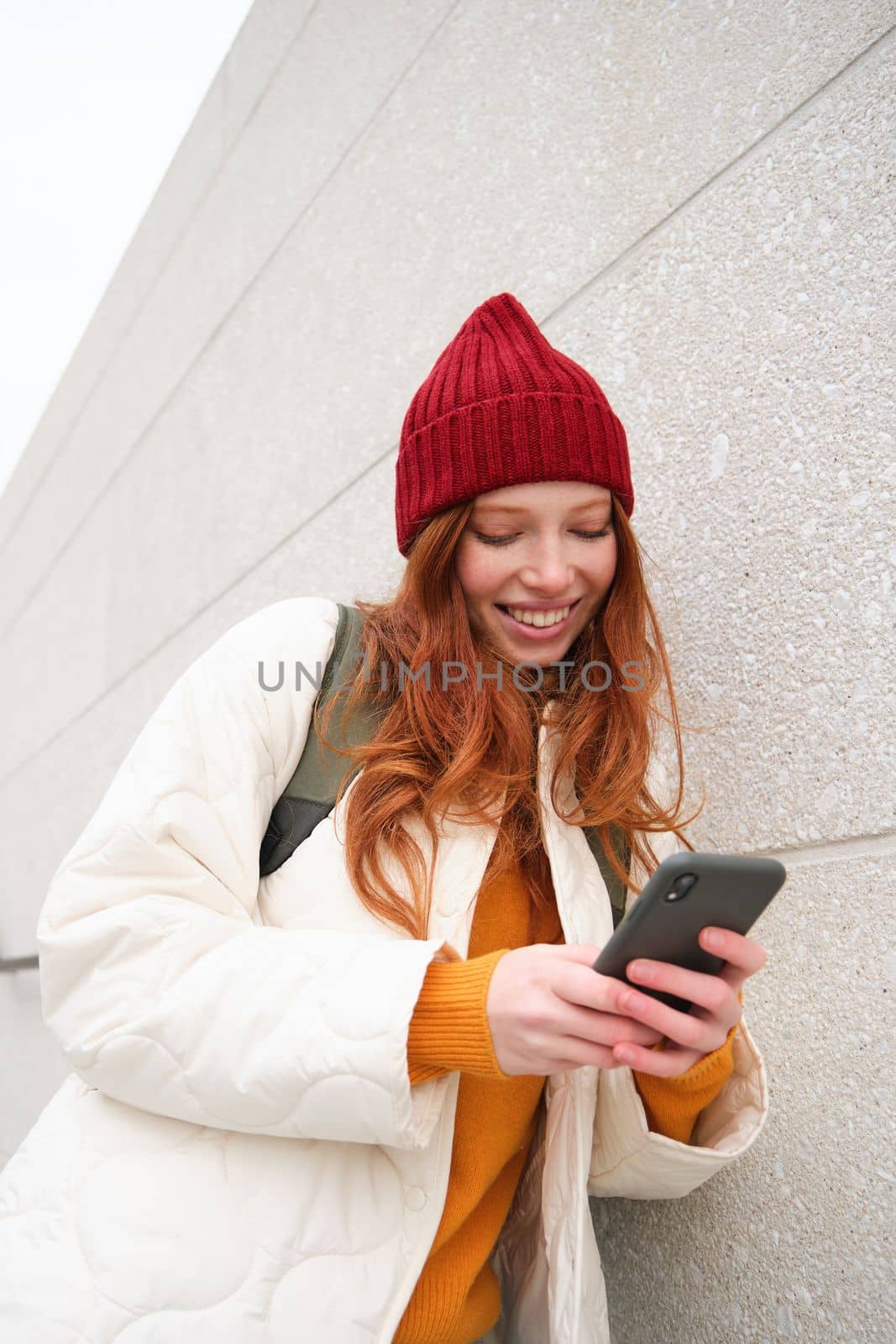 Joyful girl tourist looks at mobile phone, texts message on smartphone social media application, walks around city, looks for sightseeing on mobile app.