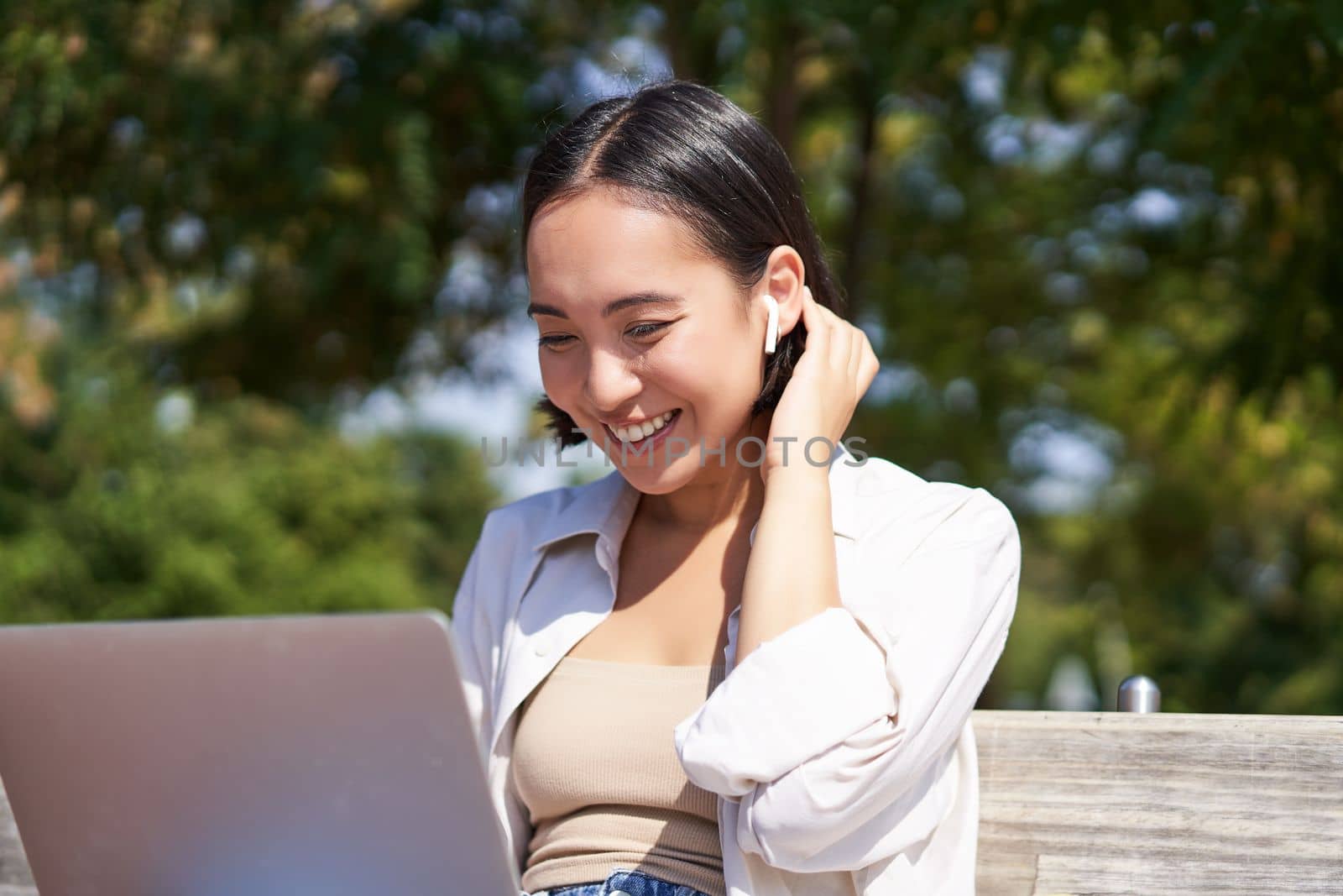 Beautiful young korean woman in wireless earphones, video chat, working outdoors in park with laptop, smiling and looking happy.