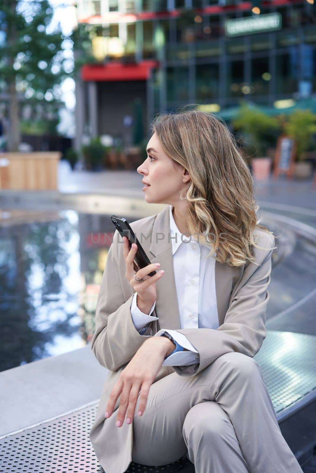 Vertical shot of businesswoman looking around, holding mobile phone, waiting at a meeting spot, sitting near fountain in city centre.