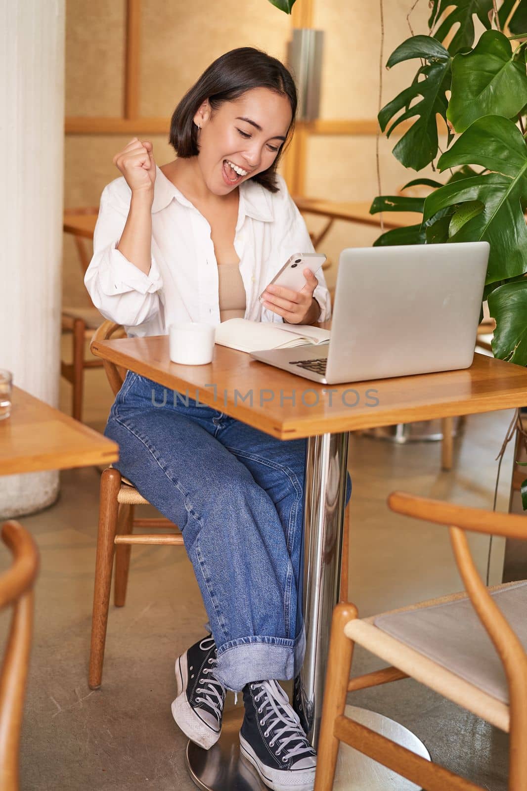 Full body shot of young asian girl sits in cafe with laptop, sees good news on smartphone and celebrates, triumphs from goal or achievement by Benzoix