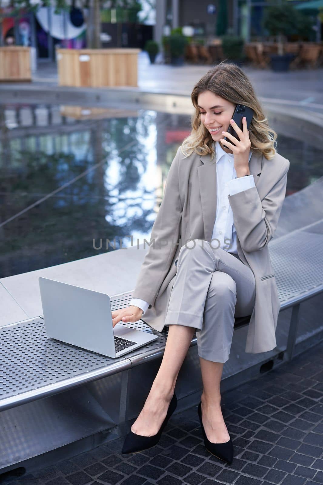 Vertical shot of corporate woman sitting outdoors with laptop, talking on mobile phone, working ouside office building while waiting for someone.