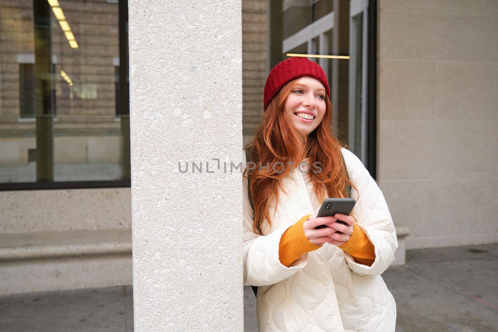 Stylish urban girl using mobile phone app, standing in city, waiting for taxi, looking at smartphone application, texting message.