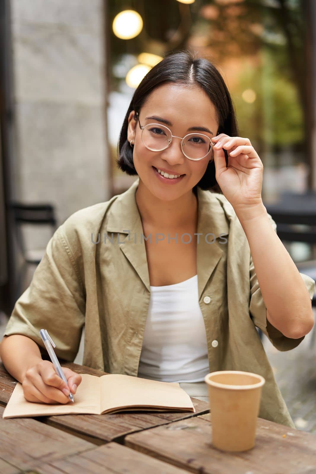 Girl studying from cafe, looking at camera while writing something in notebook, drinking coffee and smiling.