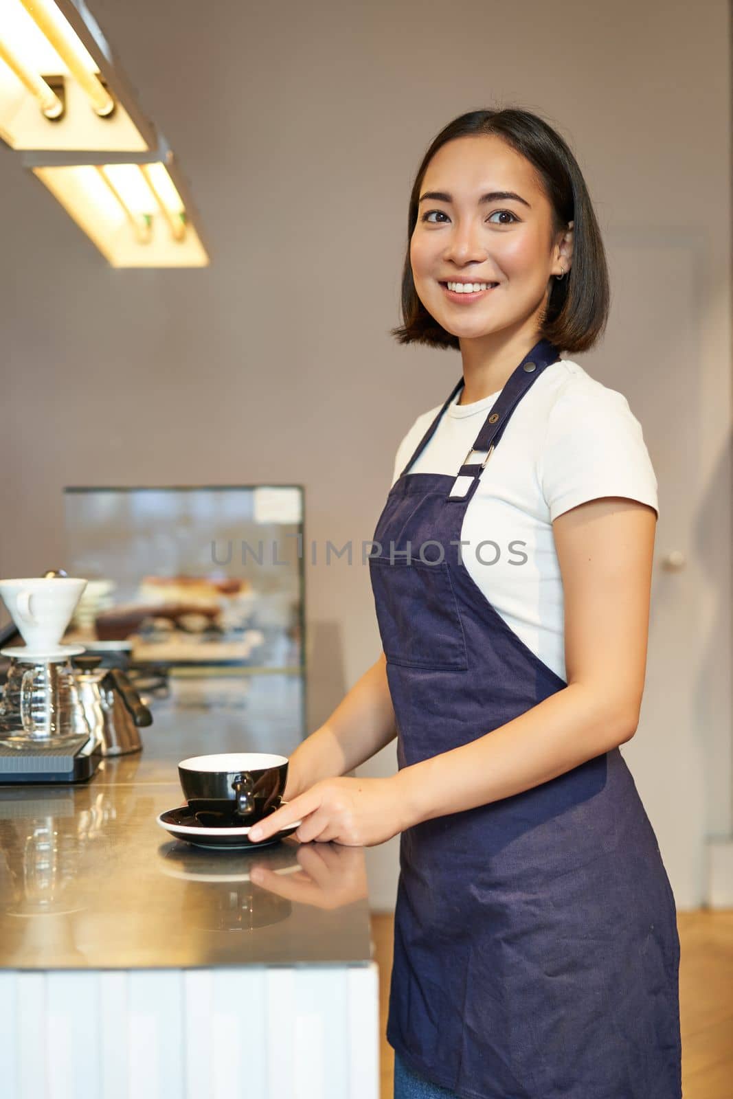 Vertical shot of smiling asian girl barista, wearing uniform, making coffee, standing near counter with cup, working in cafe by Benzoix