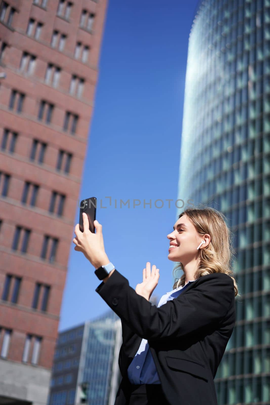 Portrait of smiling corporate woman video call on street, holding mobile phone and waving at smartphone camera, wearing suit by Benzoix