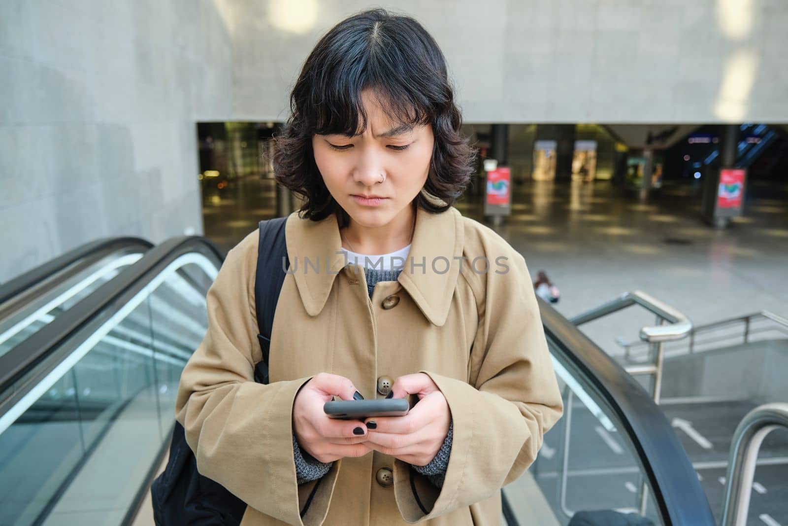 People in city. Portrait of girl looks concerned at smartphone screen with frowned worried face expression. Woman goes up escalator with mobile phone by Benzoix