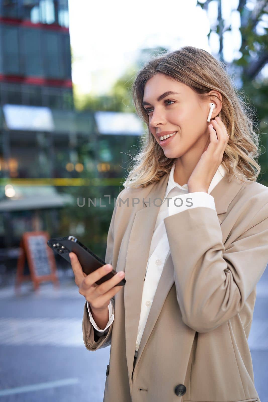 Vertical shot of businesswoman in suit, listening music in wireless headphones, holding smartphone, standing outdoors by Benzoix