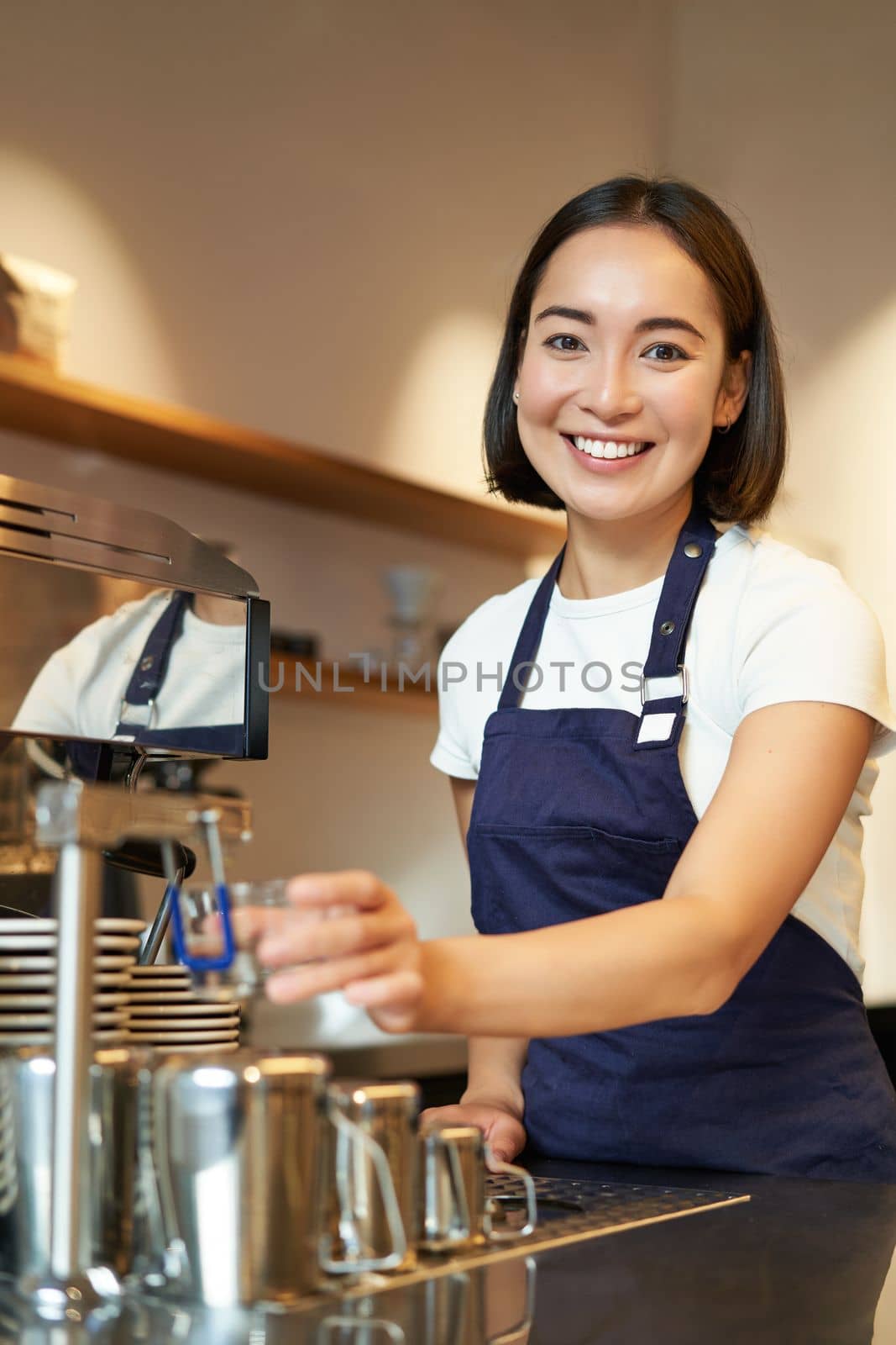 Vertical shot of young professional, smiling girl barista working in cafe, steaming milk for cappuccino drink, preparing order with coffee machine.