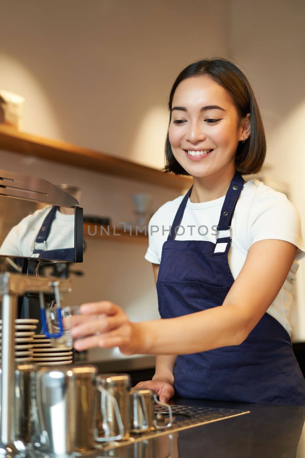 Portrait of cute barista girl working behind counter, making coffee, steaming milk for cappuccino, wearing cafe uniform blue apron.