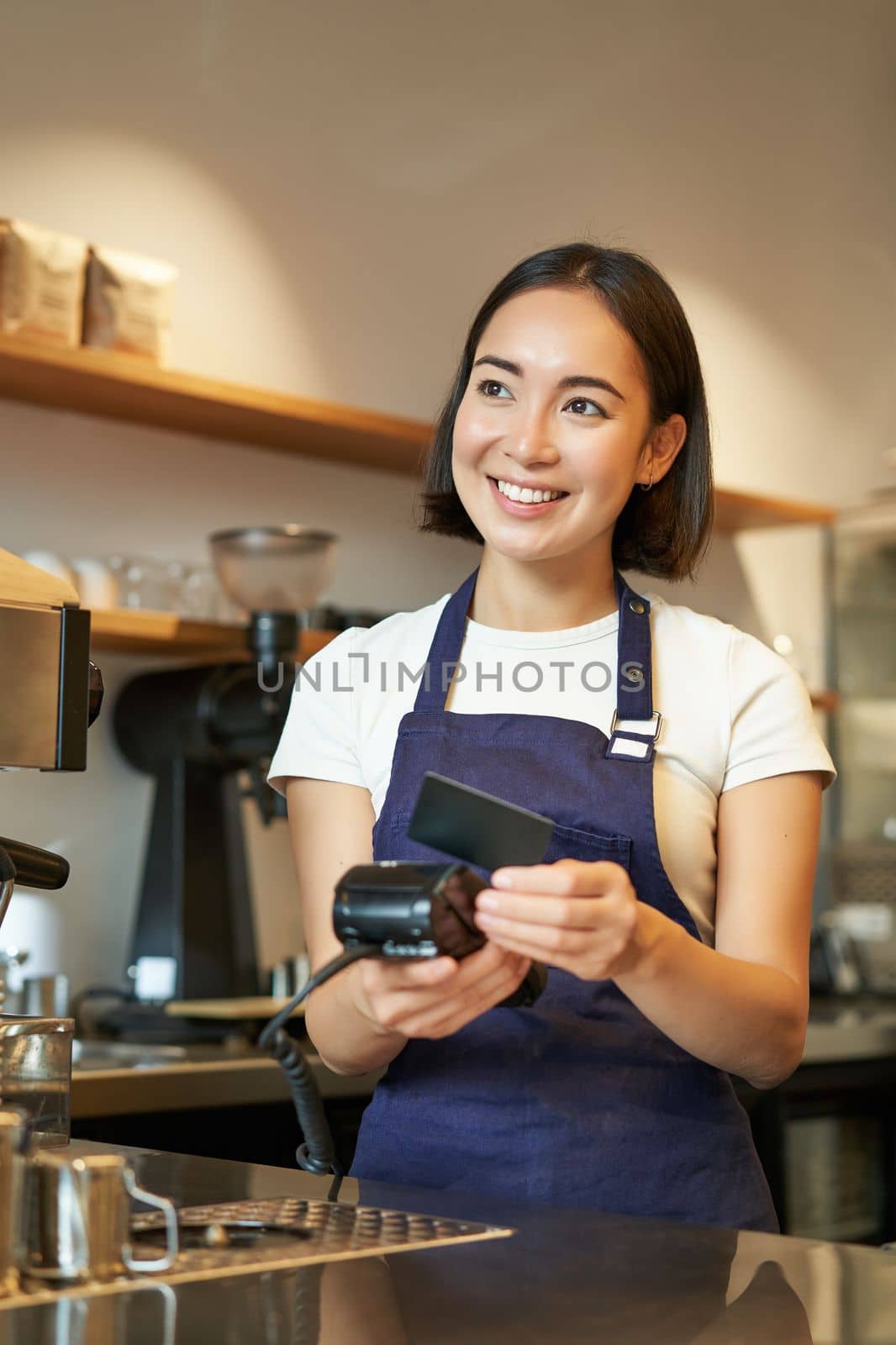 Portrait of smiling asian barista, coffee shop employee using POS terminal and credit card, helps client pay contactless in cafe.