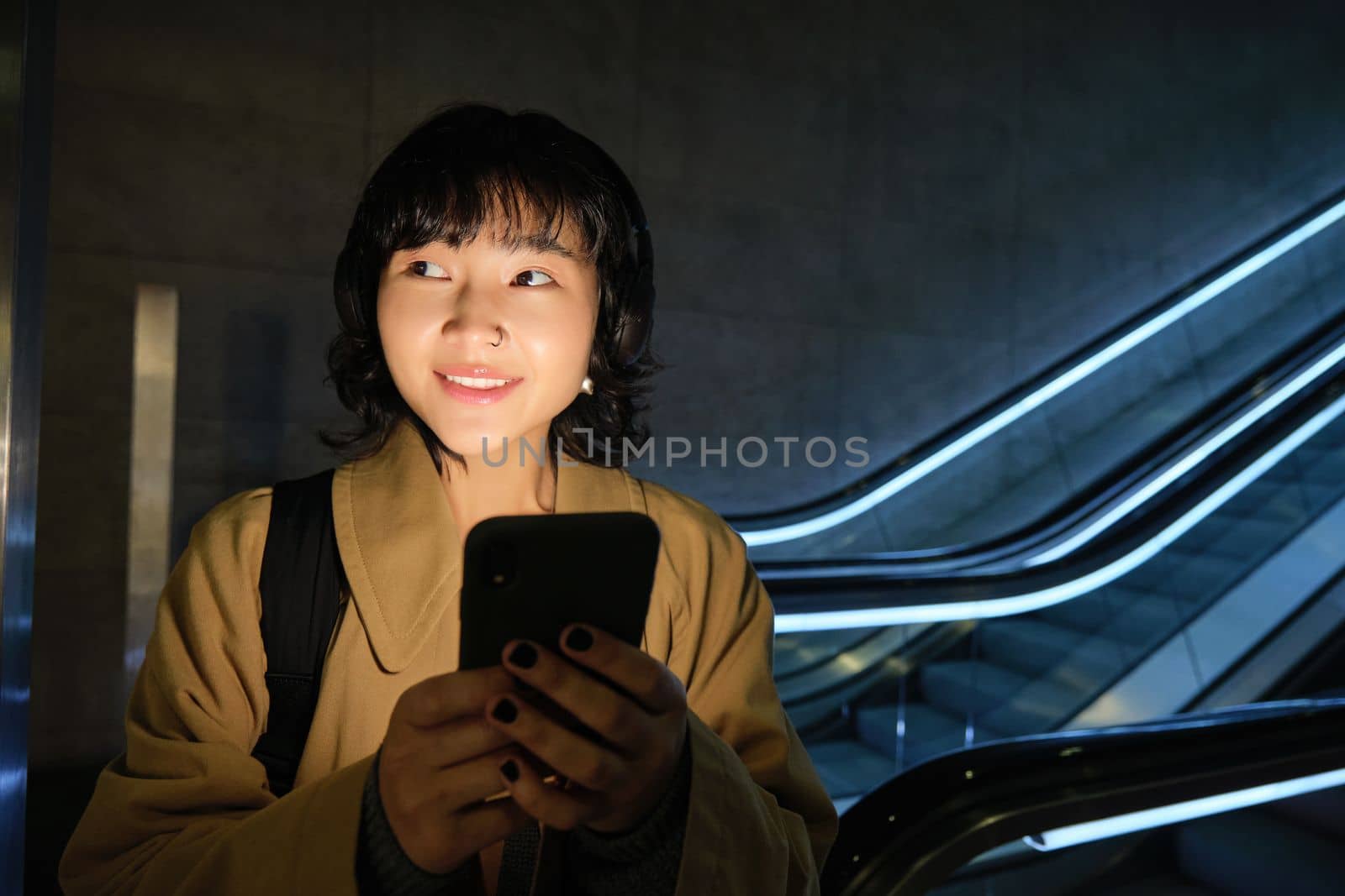 Stylish Japanese woman in headphones, standing with smartphone in hands near escalator, travelling and commuting in city, urban lifestyle concept.