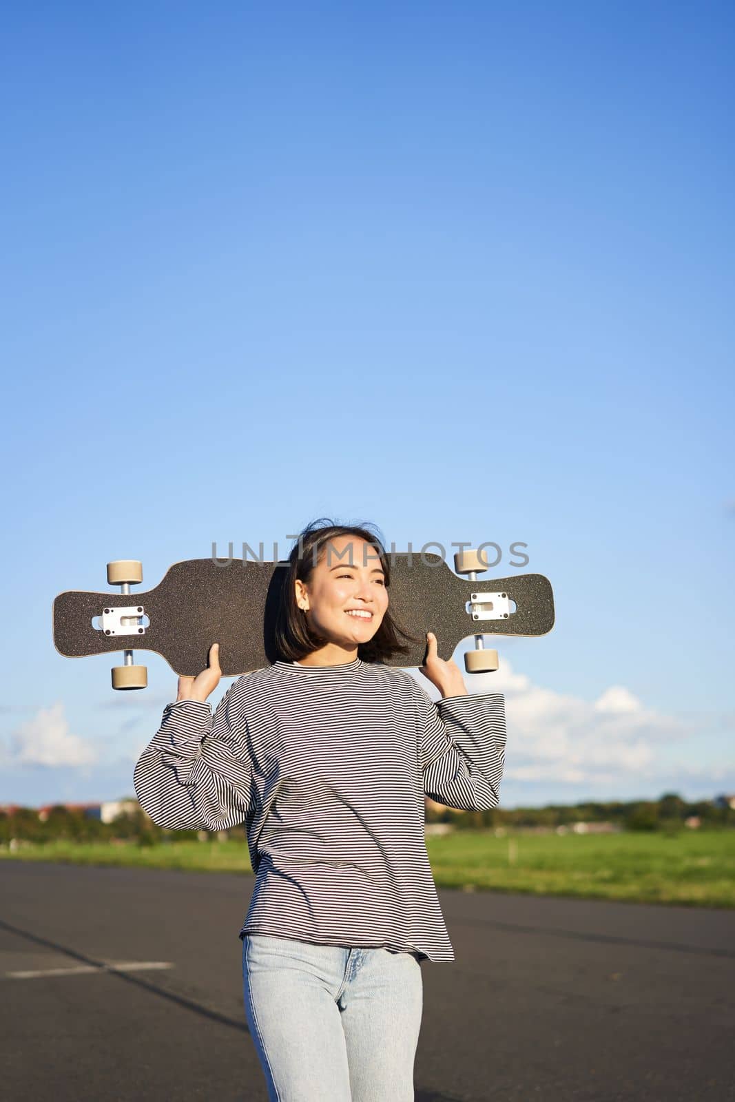 Vertical shot of carefree asian girl with longboard. Young woman skater holding cruiser on her shoulders and walking on road, skateboarding by Benzoix