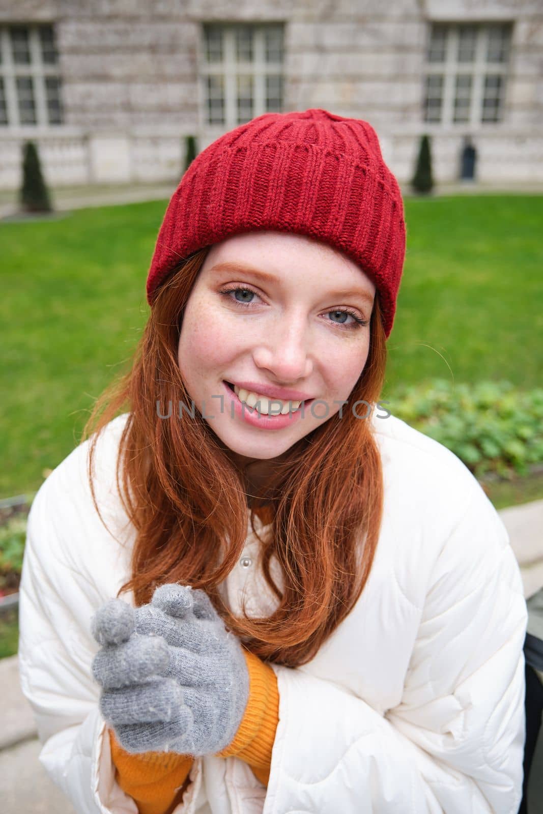 Vertical portrait of cute redhead female student in red hat and warm gloves, sits in park on bench, smiles and looks cute at camera.