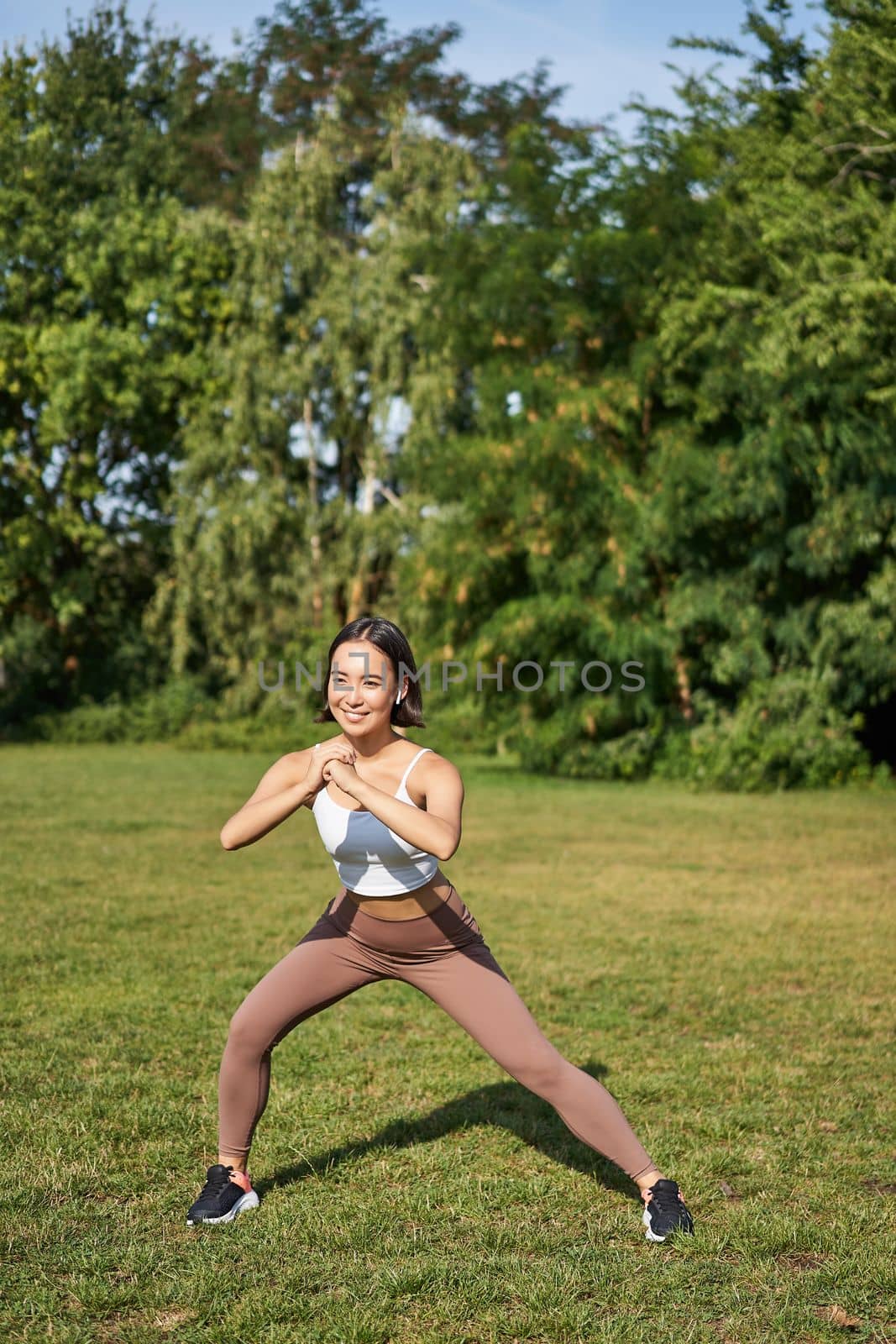 Young asian fitness girl stretching, workout in park, showing exercises, doing squats by Benzoix