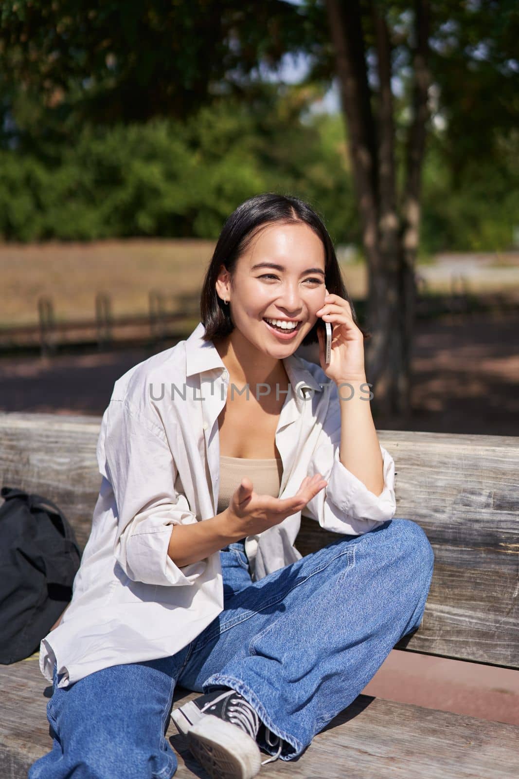 Portrait of happy young asian woman talking on mobile phone in park, sitting on bench and having a telephone call, chatting lively by Benzoix