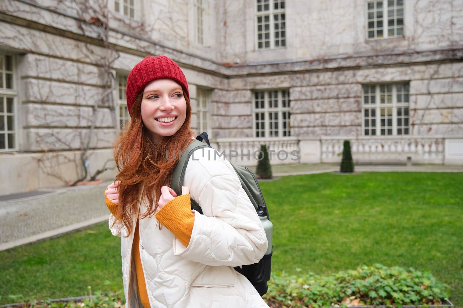 Beautiful redhead woman with backpack, tourist walking around city park and looks around, wears red hat and warm coat.