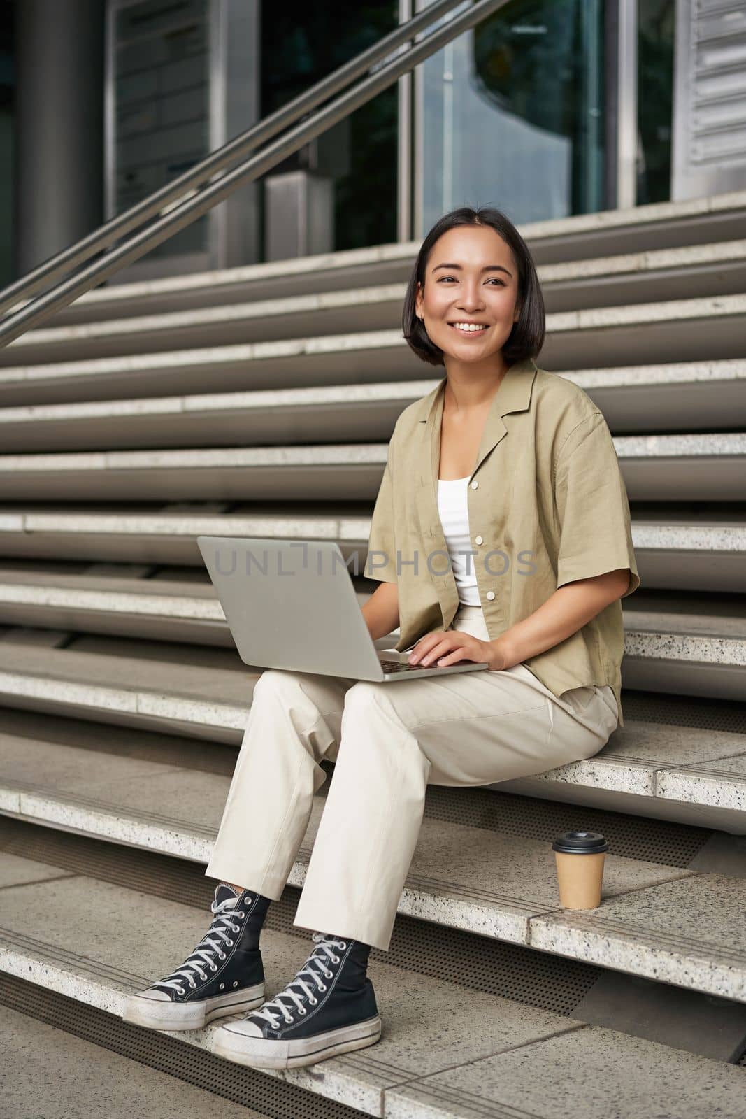 Vertical shot of asian girl sits with laptop, drinks coffee on university stairs. Young woman, student does her homework outdoors by Benzoix