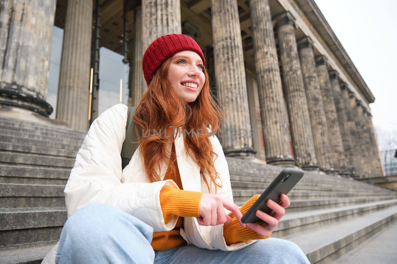 Portrait of young urban girl in red hat, sits on stairs near museum, holds mobile phone, connects to public wifi and surfs net, uses smartphone apps by Benzoix