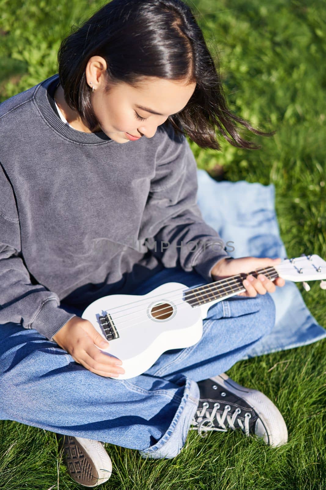 Vertical shot of girl musician, looking with care at her white ukulele guitar, playing music in park, sitting on grass.