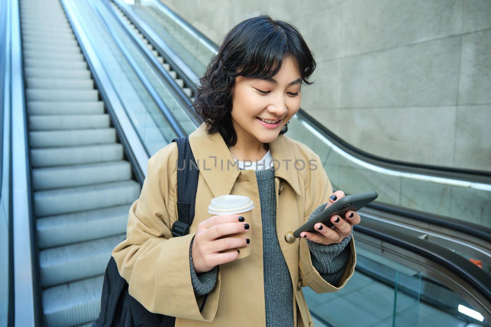 Young stylish girl with cup of coffee, drinks cappuccino to go, goes down escalator and looks at mobile phone, chats, reads message on smartphone by Benzoix