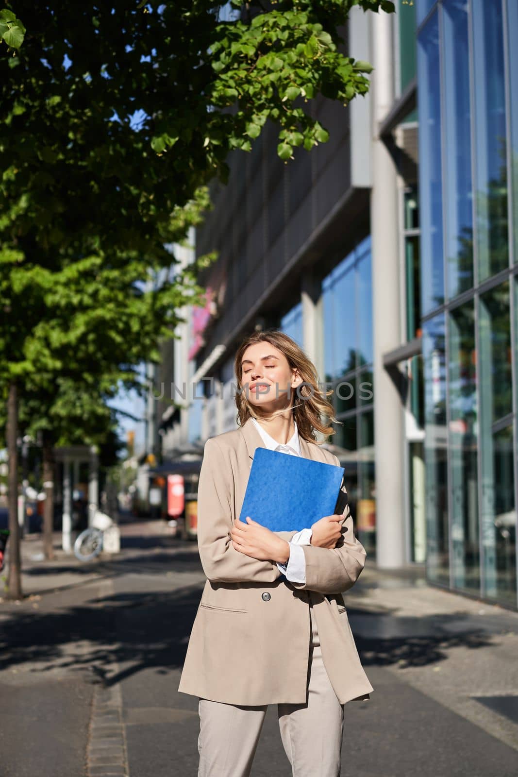 Vertical shot of businesswoman with folder, standing on a street near office buildings, wearing suit for office work by Benzoix