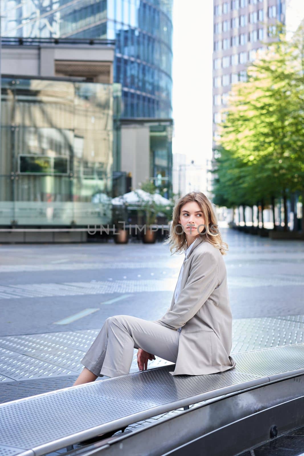 Female corporate worker sitting outdoors near office buildings, turn back and look behind, waiting for coworker on lunch break.