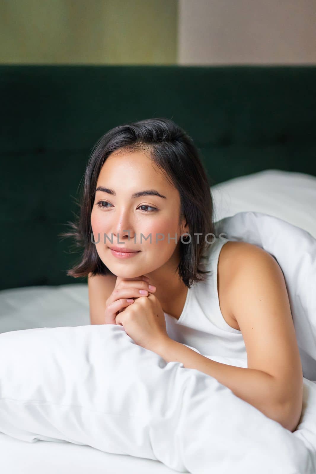 Vertical shot of beautiful asian woman lying in her bed on pillow, covered in duvet, looking thoughtful and smiling, waking up and thinking.