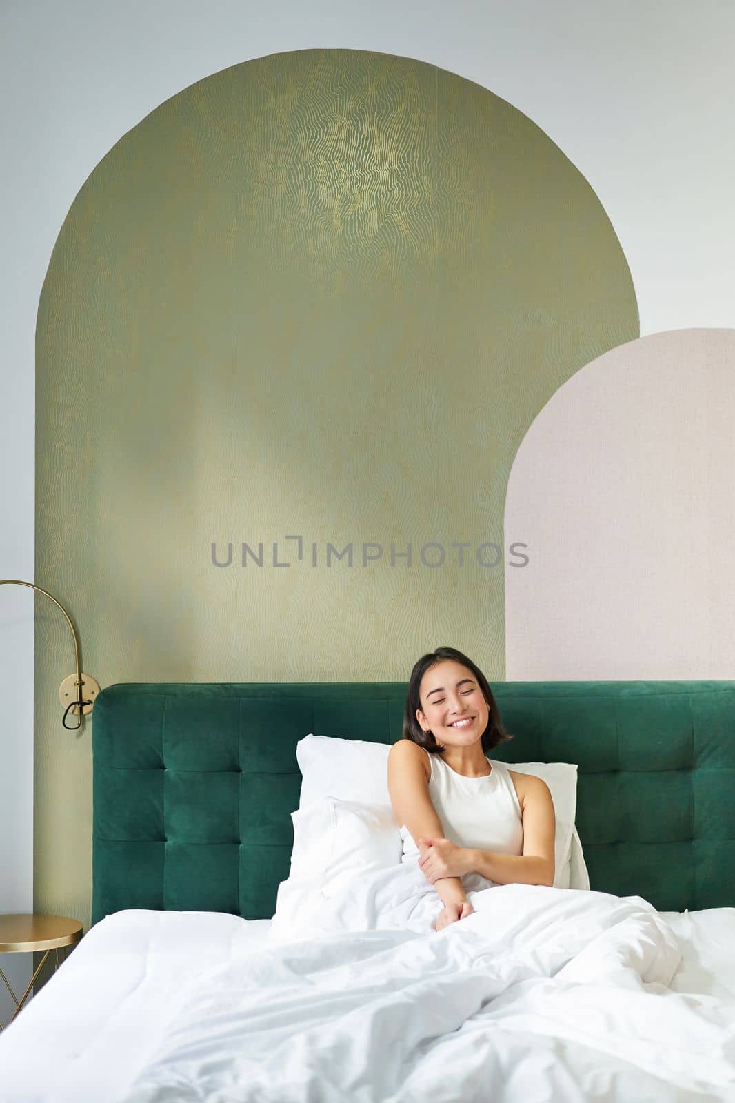 Vertical shot of beautiful asian girl lying in bedroom with sophisticated interrior, smiling and looking happy in morning, spending time in bed.