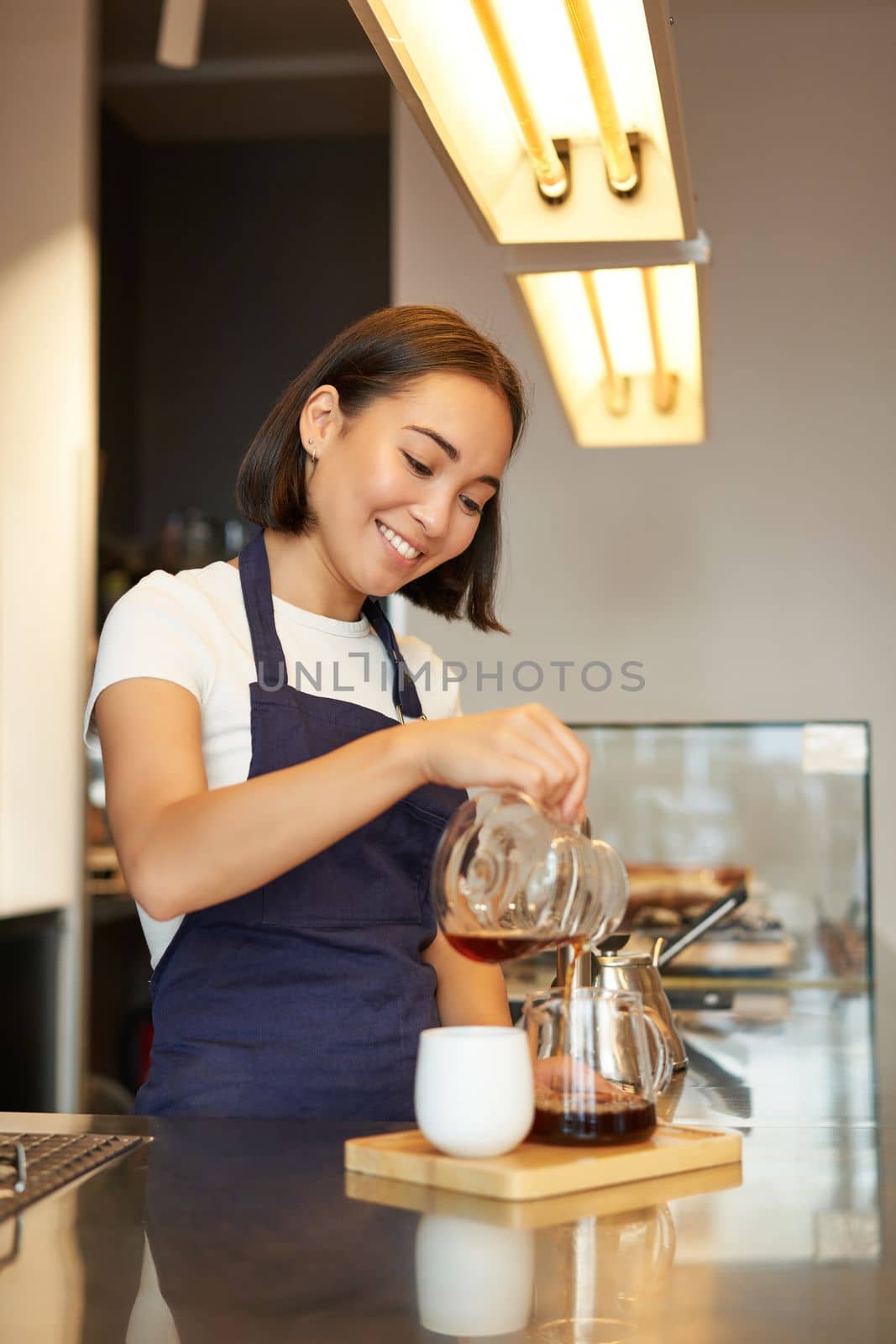 Vertical shot of smiling girl working as barista, prepare pour over, making filter coffee batch brew, standing at counter in cafe in apron.