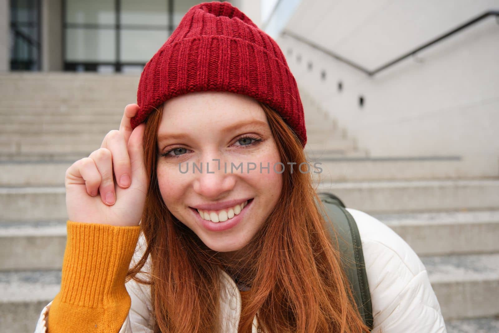 Stylish redhead girl in warm red hat, smiling relaxed, sitting with backpack on stairs near building, waits for someone outdoors by Benzoix