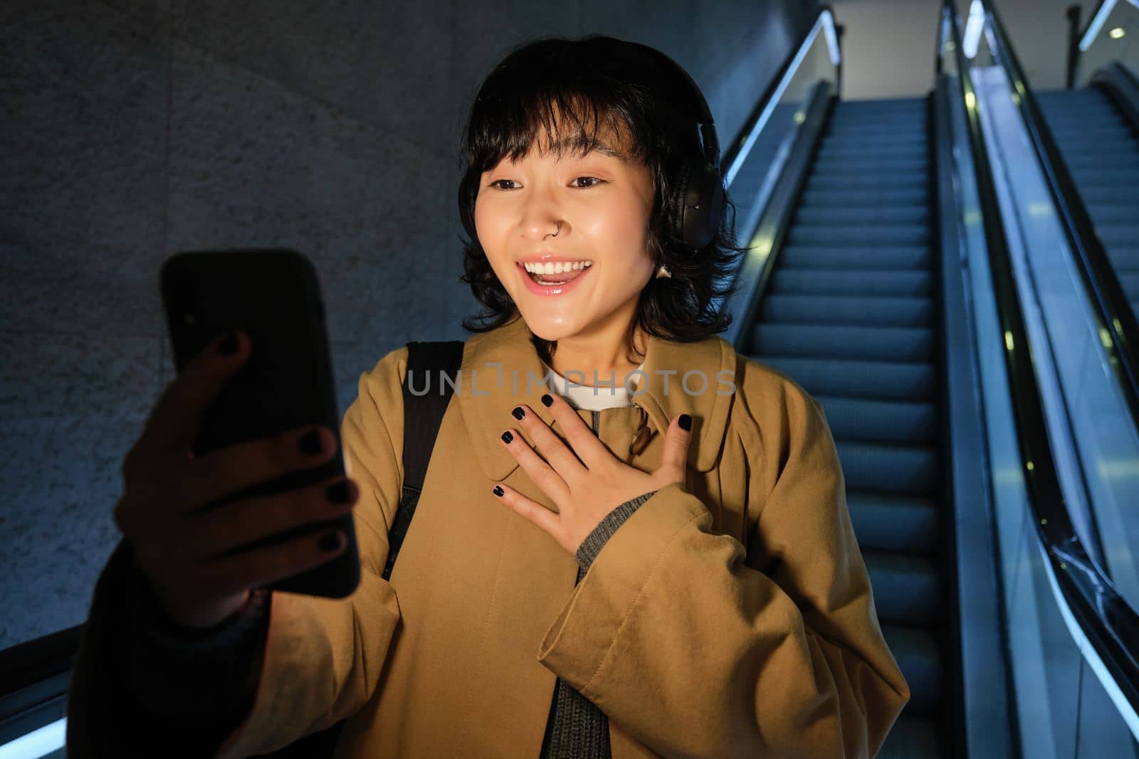 Surprised asian girl looks at smartphone screen, feeling amazed by smth she read on mobile phone, going down escalator in city, commuting to university.