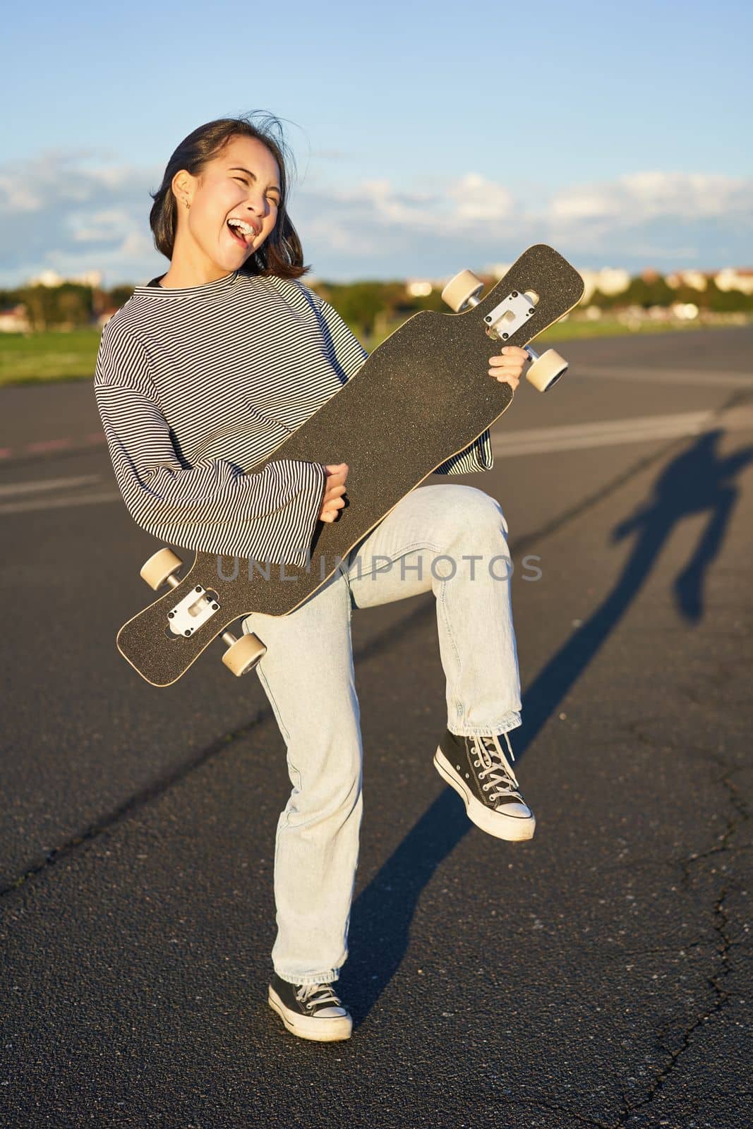 Funny asian girl enjoying skating, holding skateboard like guitar and shadow playing, having fun outdoors by Benzoix