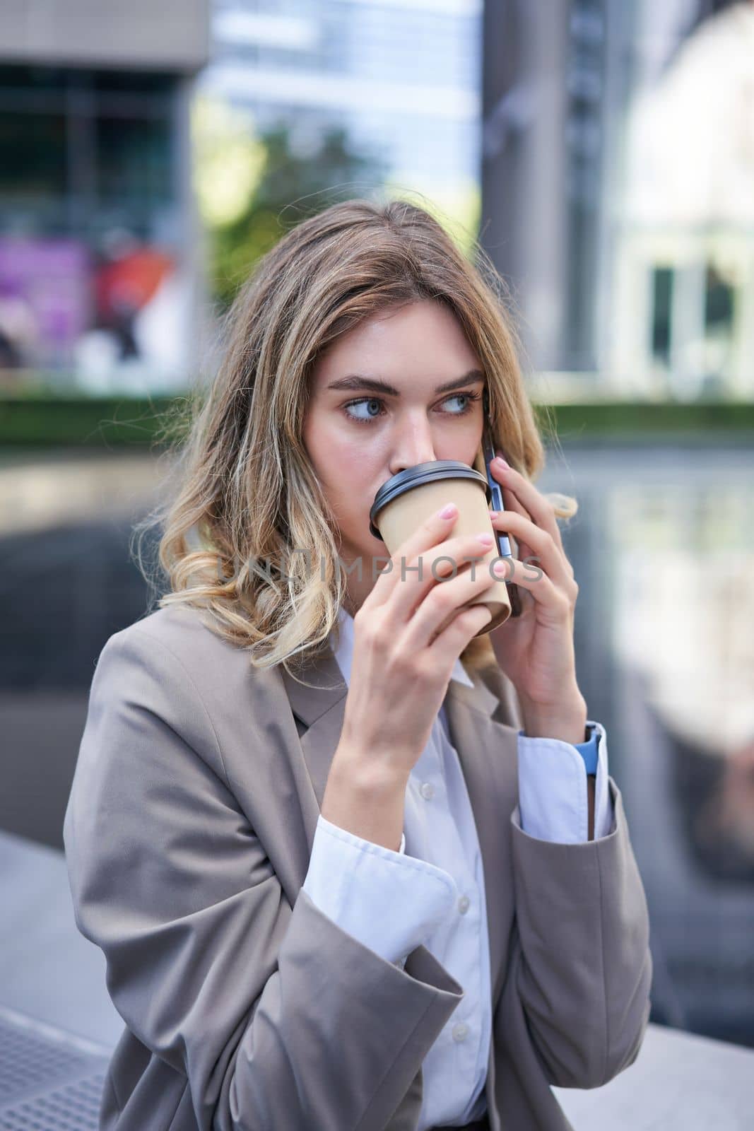 Portrait of beautiful woman talking on mobile phone and drinking coffee. Businesswoman drinks takeaway in city centre, calling someone by Benzoix