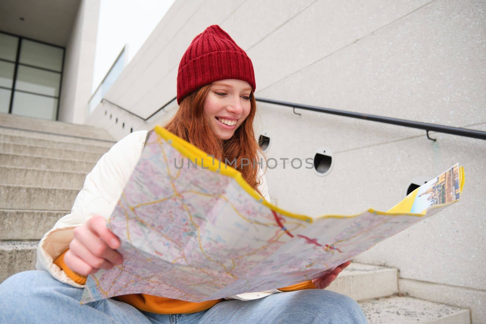 Beautiful girl tourist sits on stairs with city map, plans her journey, looks for direction while travelling around town, searches route for sightseeing.