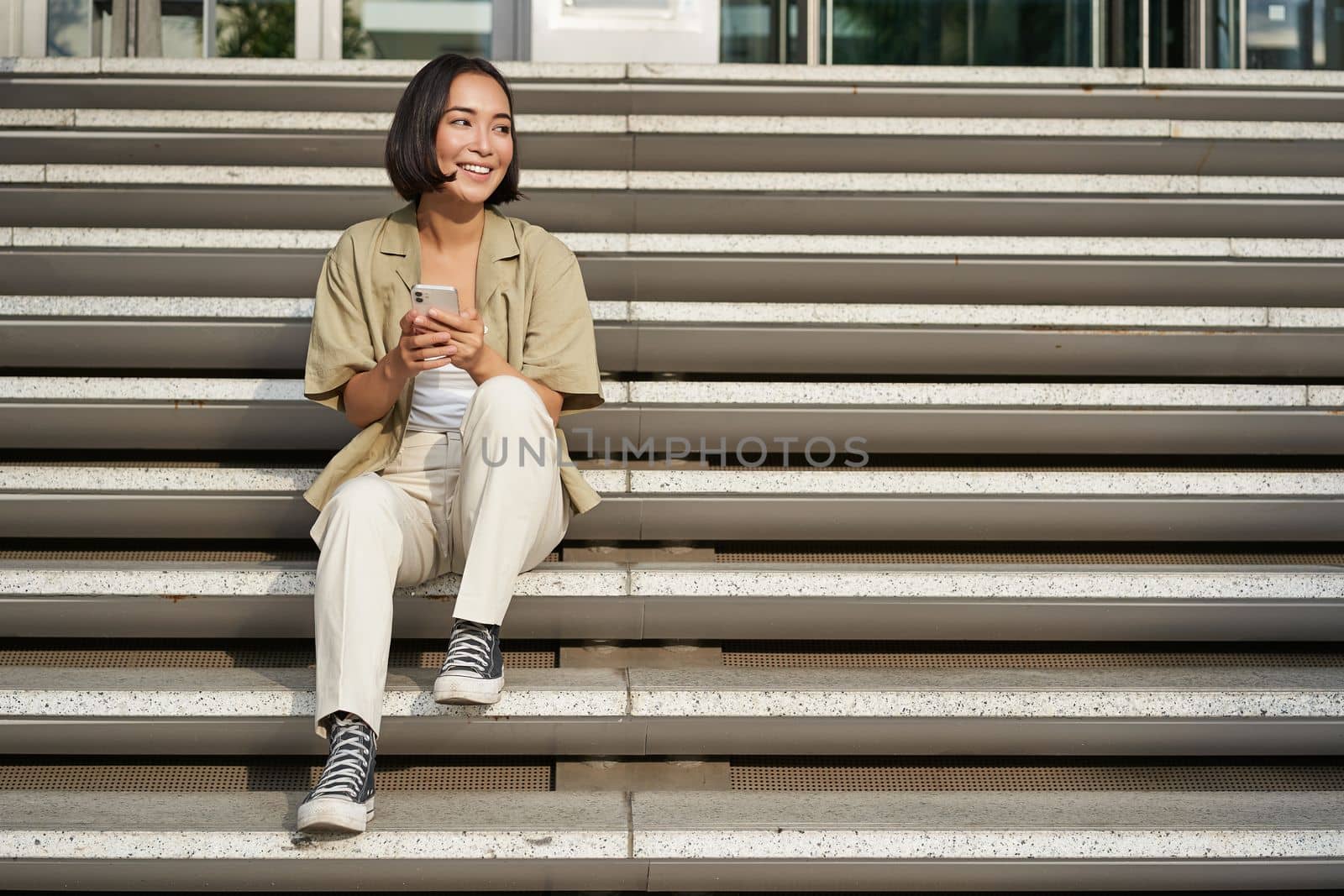 Portrait of smiling asian girl sits on stairs with her smartphone, browsing internet on mobile phone, resting outdoors in city centre.