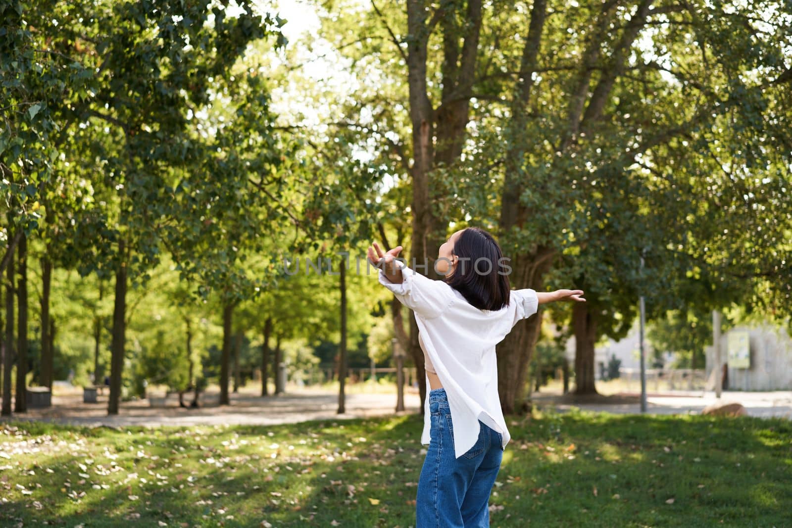 Carefree asian girl dancing, feeling happiness and joy, enjoying the sun on summer day, walking in park with green trees.