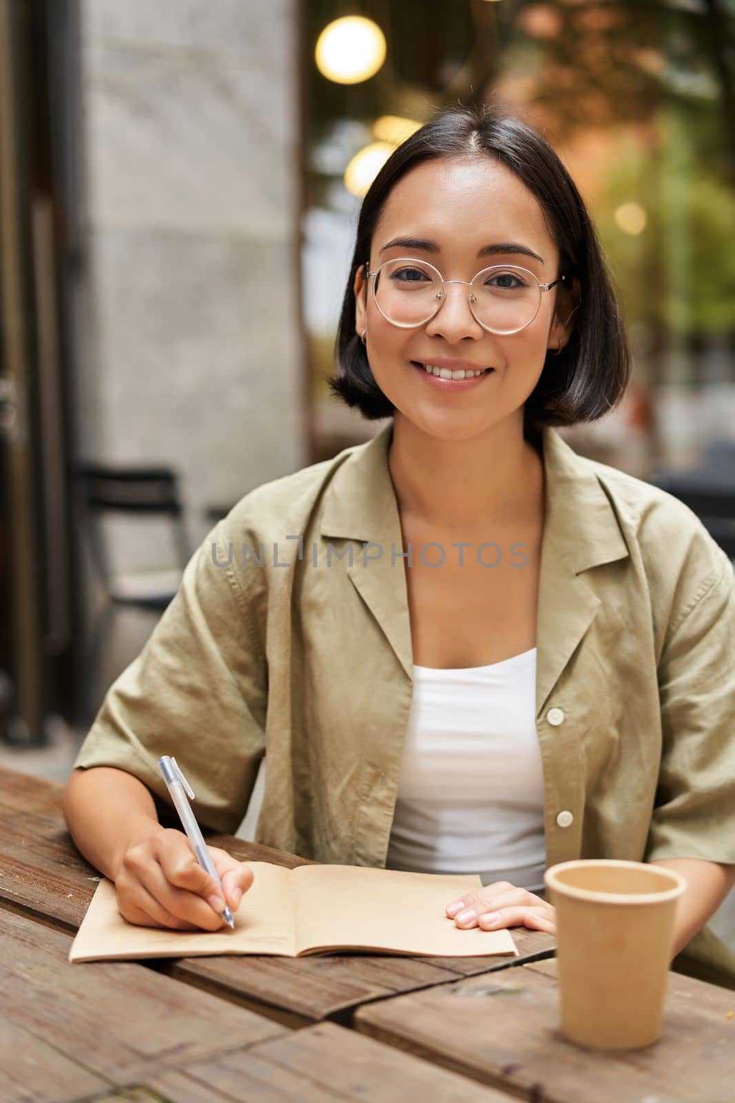 Vertical shot of young asian student, girl in glasses holding pen, making notes, writing in notebook and drinking coffee in cafe.