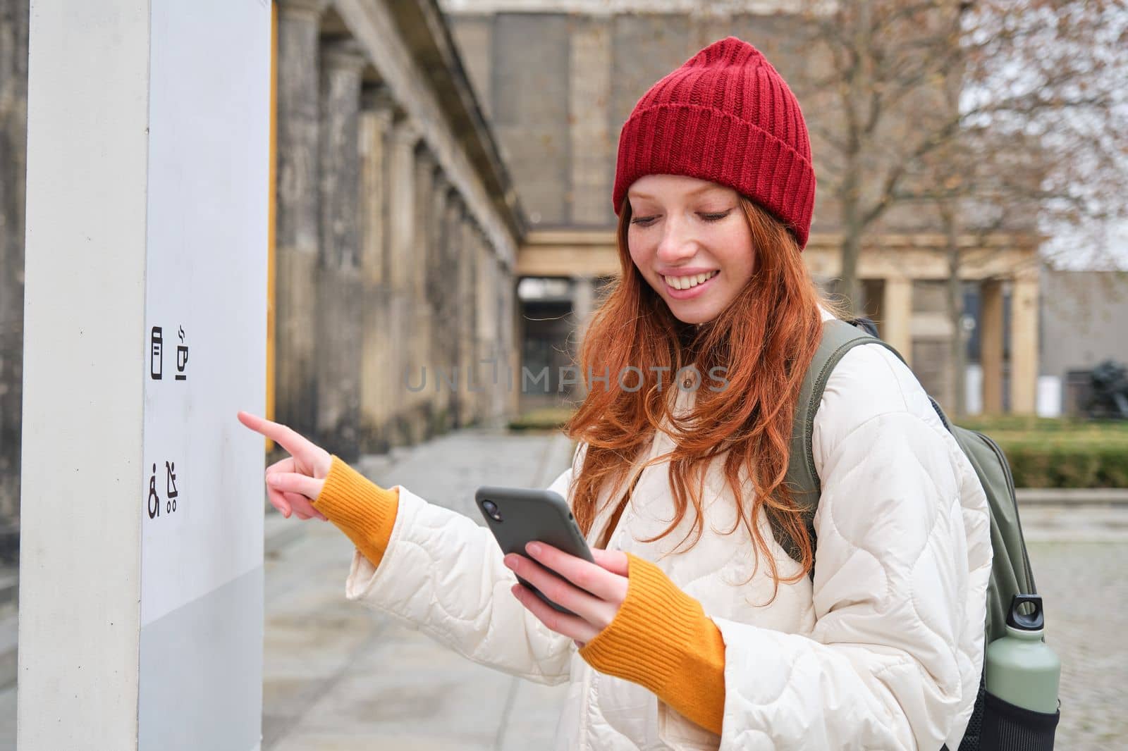 Smiling redhead girl tourist, walks around city and explores popular landmarks, sightseeing, holding smartphone, looking at her mobile app and checks with city map.