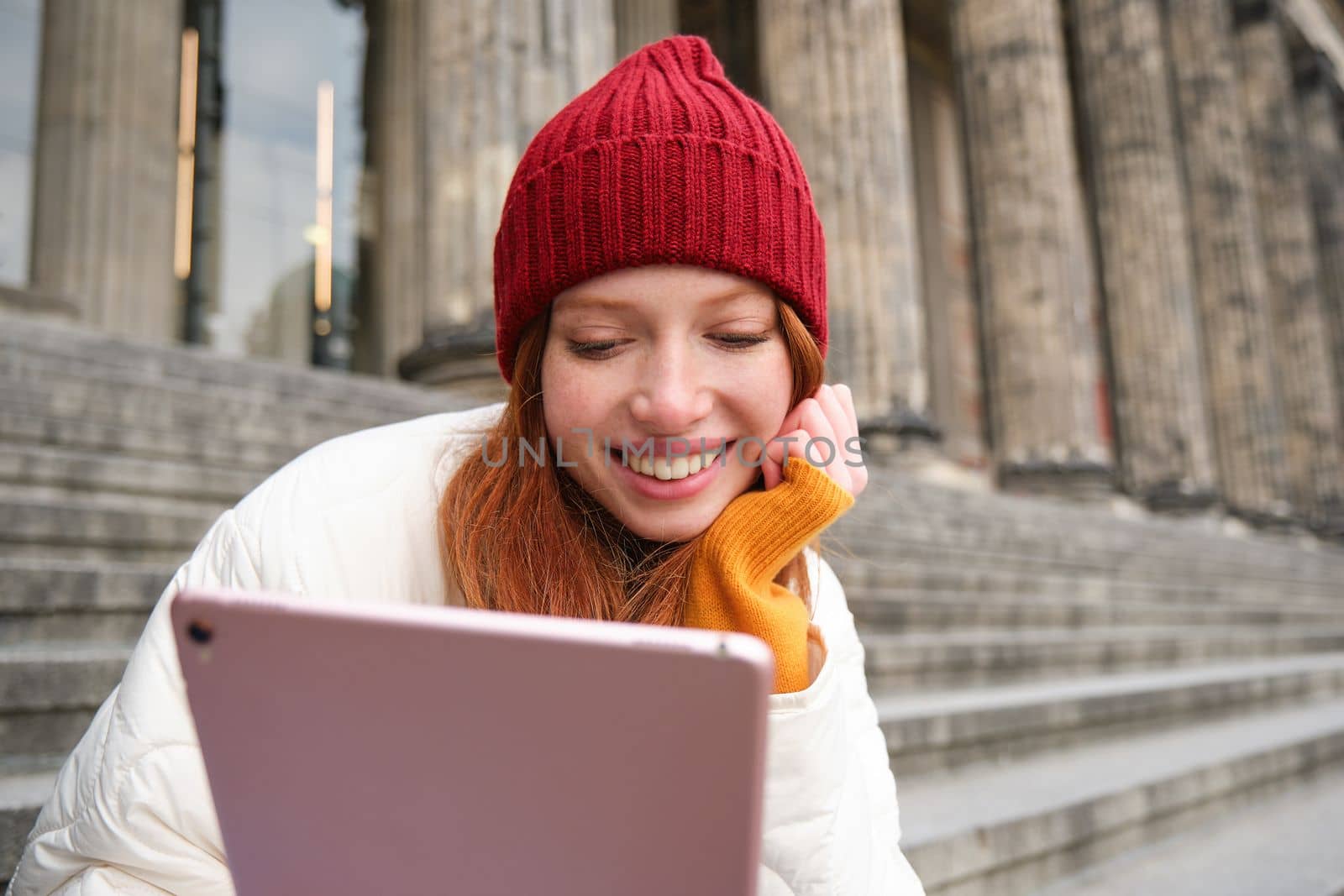 Portrait of young redhead woman sitting outdoors on stairs, reading e-book on digital tablet, wearing red hat and warm clothes by Benzoix